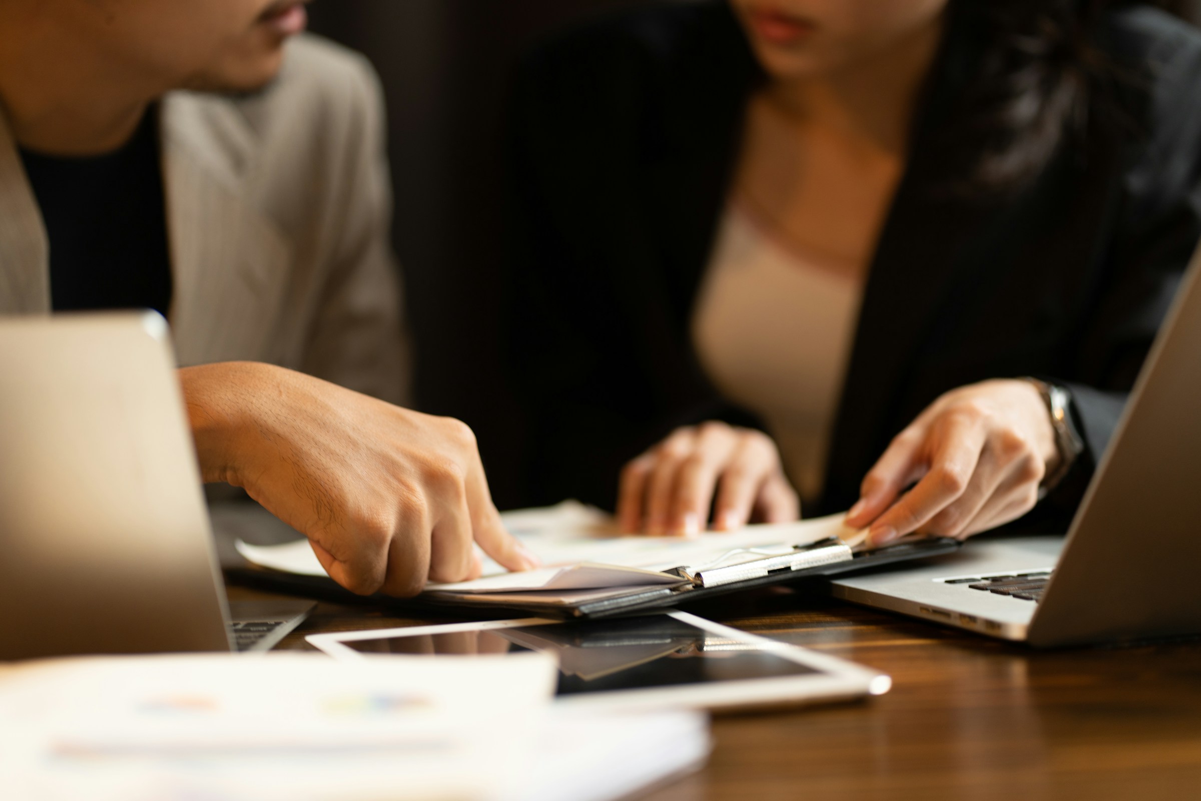 Two individuals are sitting at a table with laptops and papers, discussing and pointing at documents, possibly deliberating on what is required to obtain a Tax Compliance Certificate.