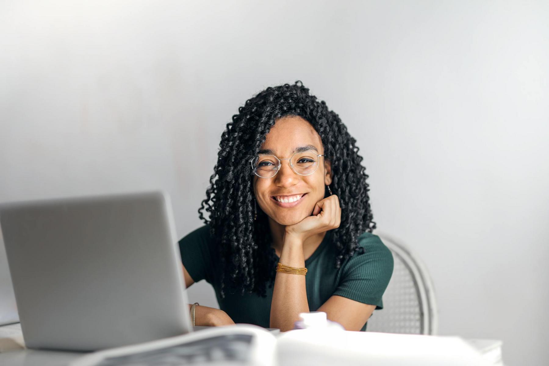 Woman with curly hair and glasses smiling at a desk with a laptop and open book in a bright room.