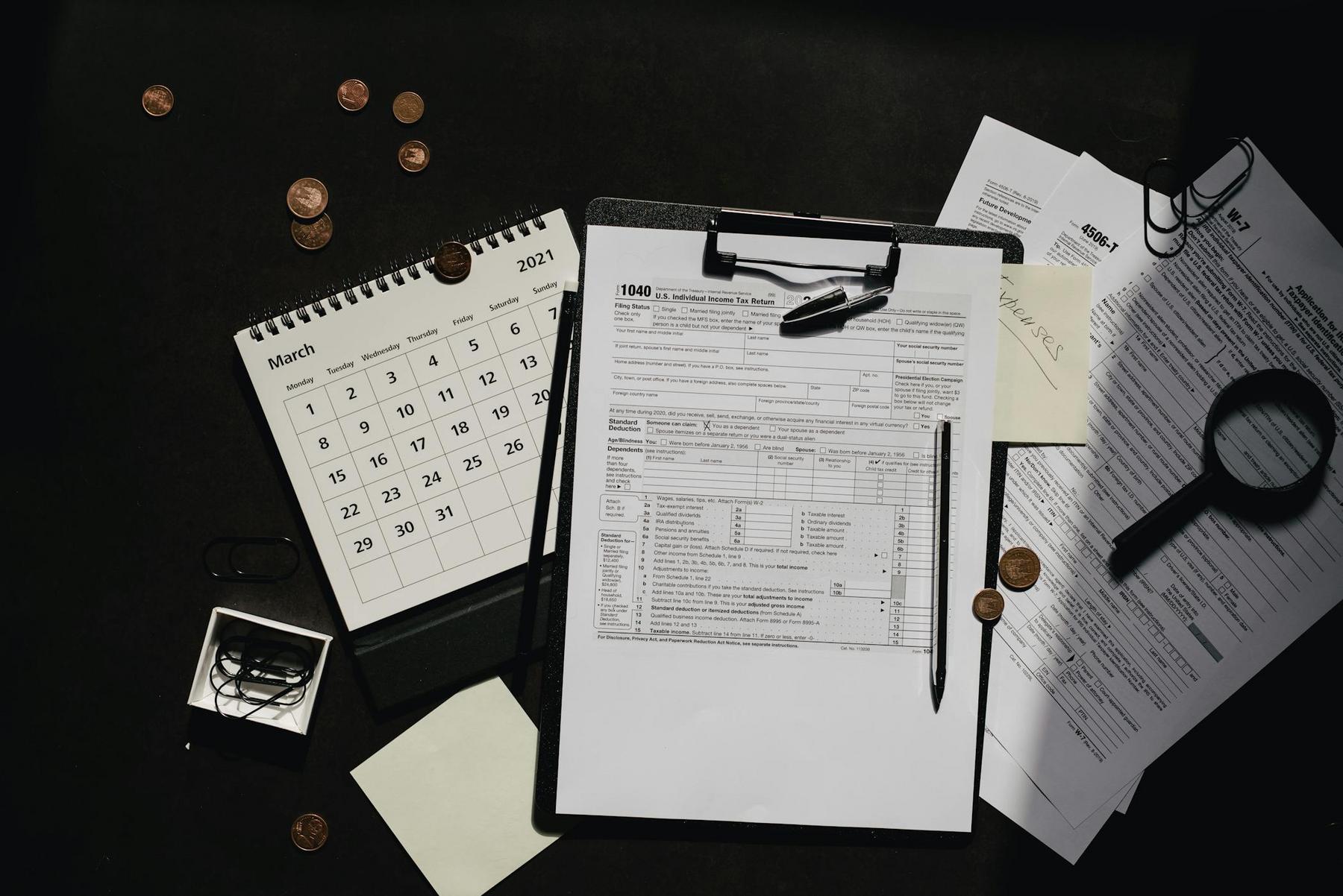 A clipboard with a 1040 tax form, calendar, magnifying glass, documents, eyeglasses, coins, paper clips, and sticky notes arranged on a dark surface.