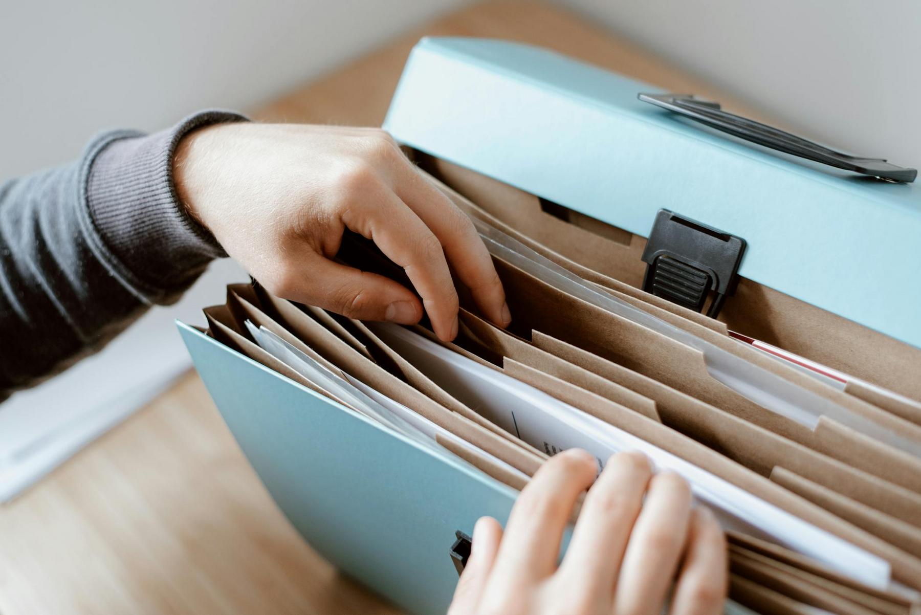 A person’s hands organizing papers in different sections of a blue accordion file folder on a wooden surface.
