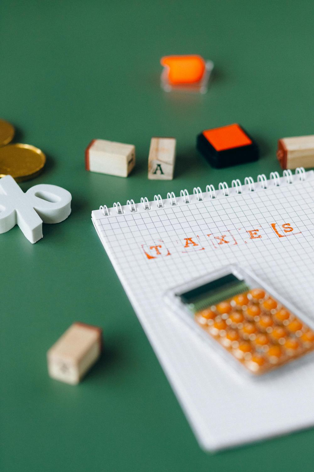 A notebook with "TAXES" stamped on squared paper, a calculator, rubber stamps, coins, and a pair of white scissors on a green surface.