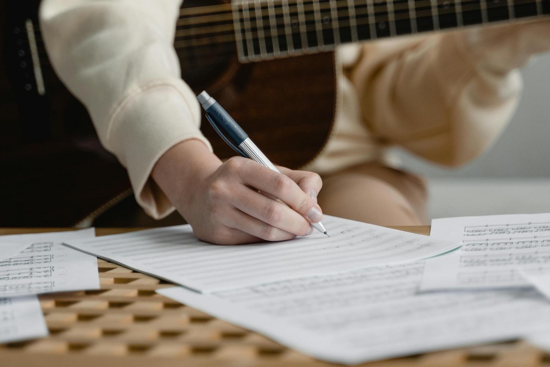 A person writes music on sheet paper with a pen, while holding a guitar. Several sheets of music notation are spread out on a wooden table.