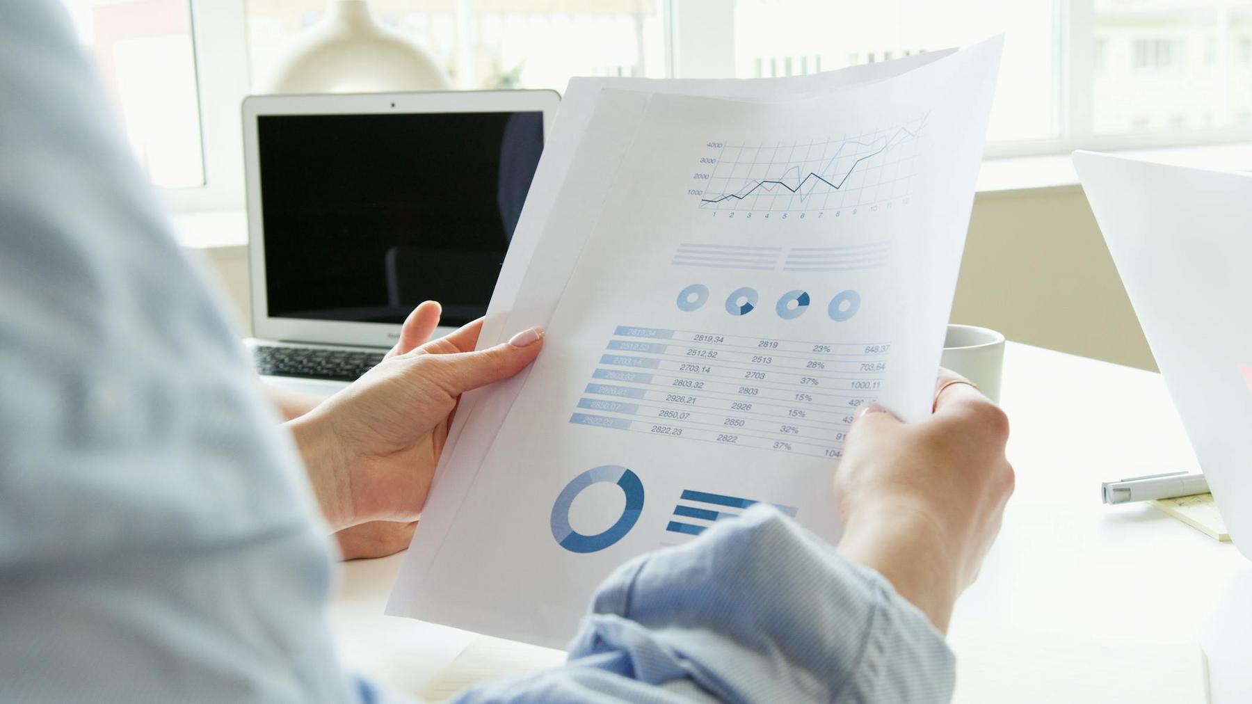 Person holding printed business charts and graphs at a desk, with a laptop and cup in the background.