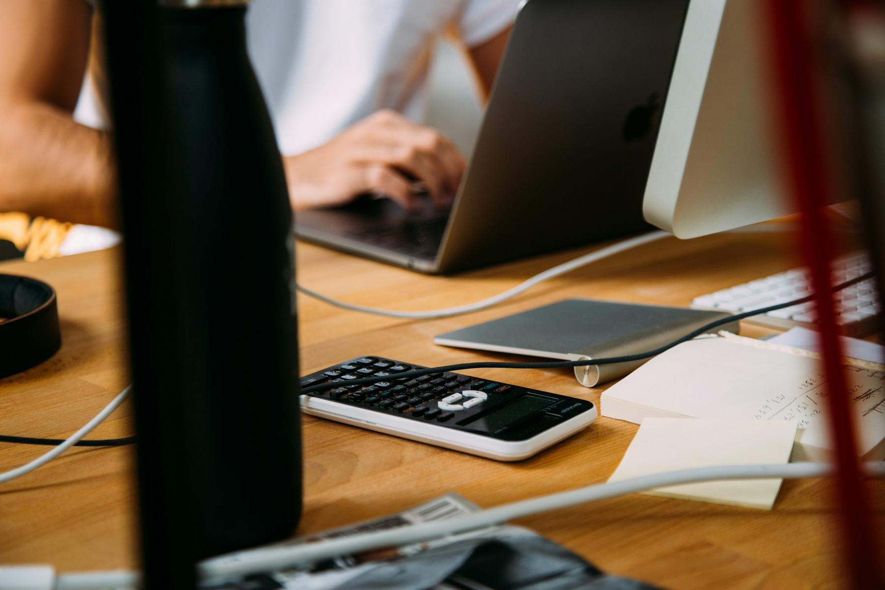 A person types on a laptop at a cluttered desk, with a calculator, documents, and an iMac nearby.