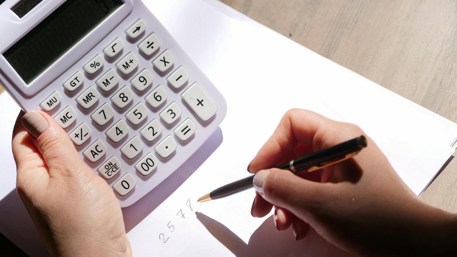 A person holds a calculator in one hand and writes numbers on a sheet of white paper with a pen using the other hand.