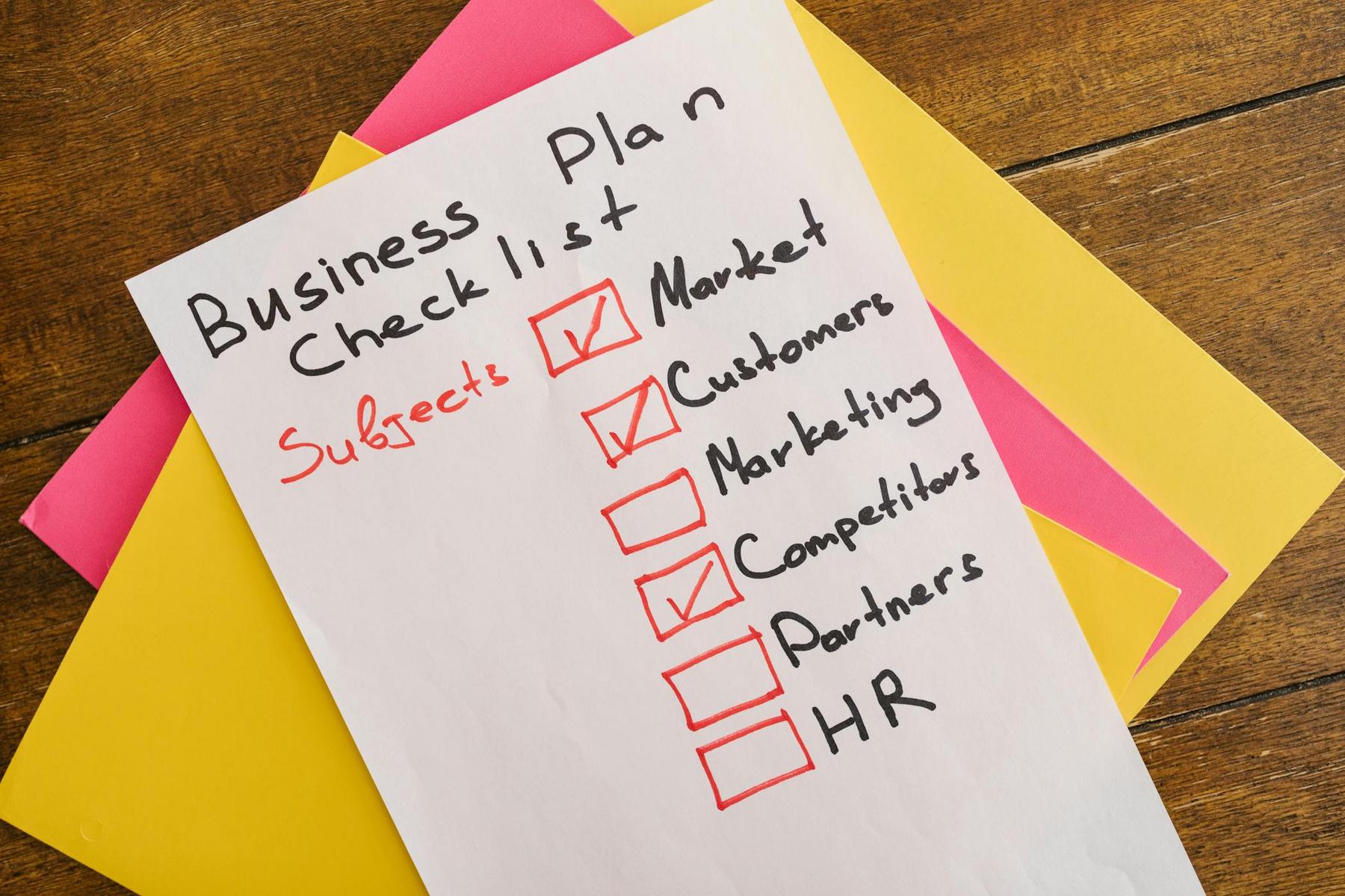A handwritten business plan checklist on white paper lists Market, Customers, Marketing, Competitors, Partners, and HR, with the first three items checked. The sheet rests on yellow and pink folders.