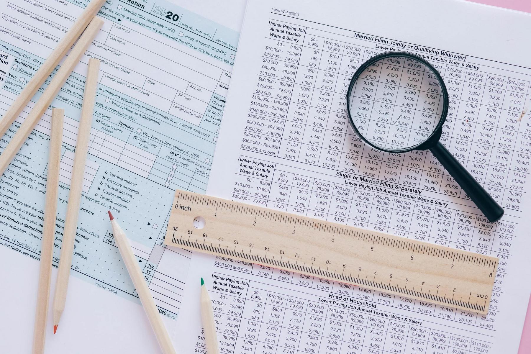 A magnifying glass, ruler, and pencils are placed on top of tax forms and a tax table.