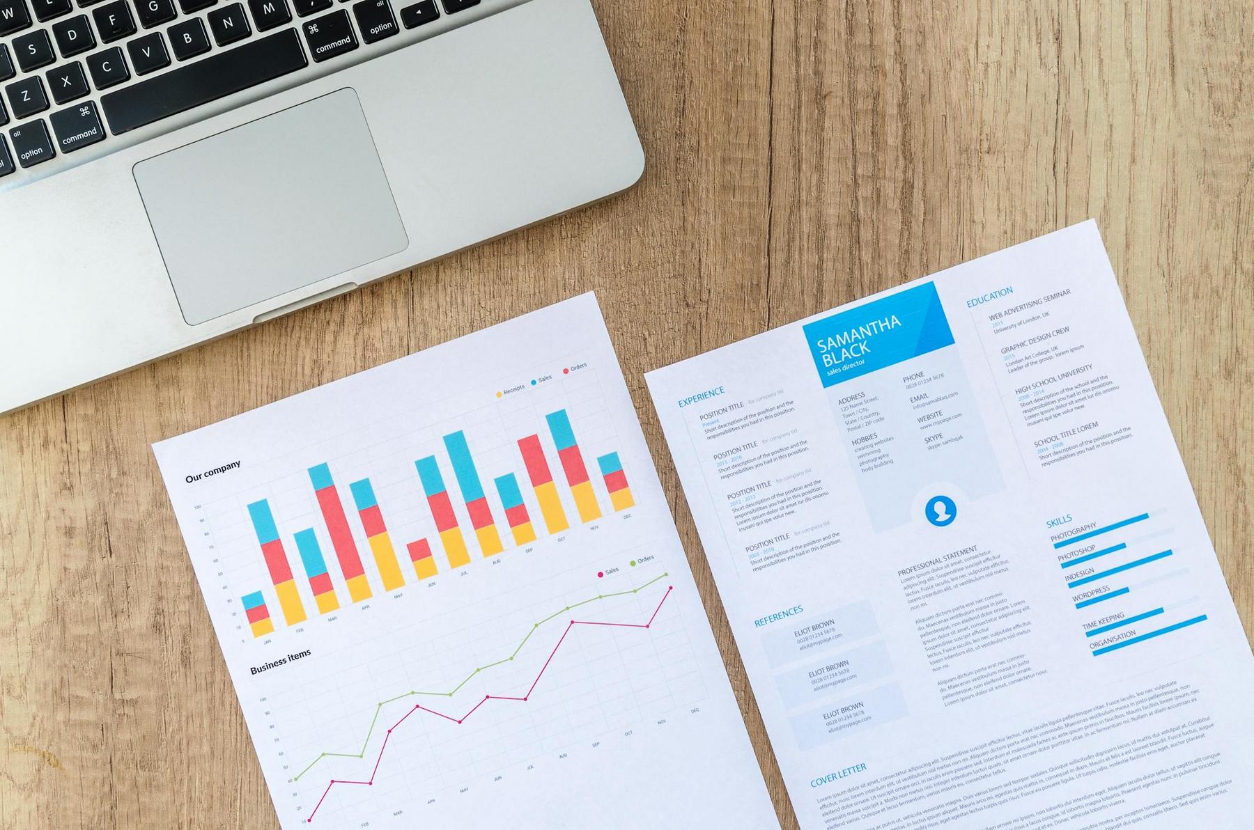 A laptop and two sheets of paper on a wooden desk; one has colorful charts, the other appears to be a professional resume.
