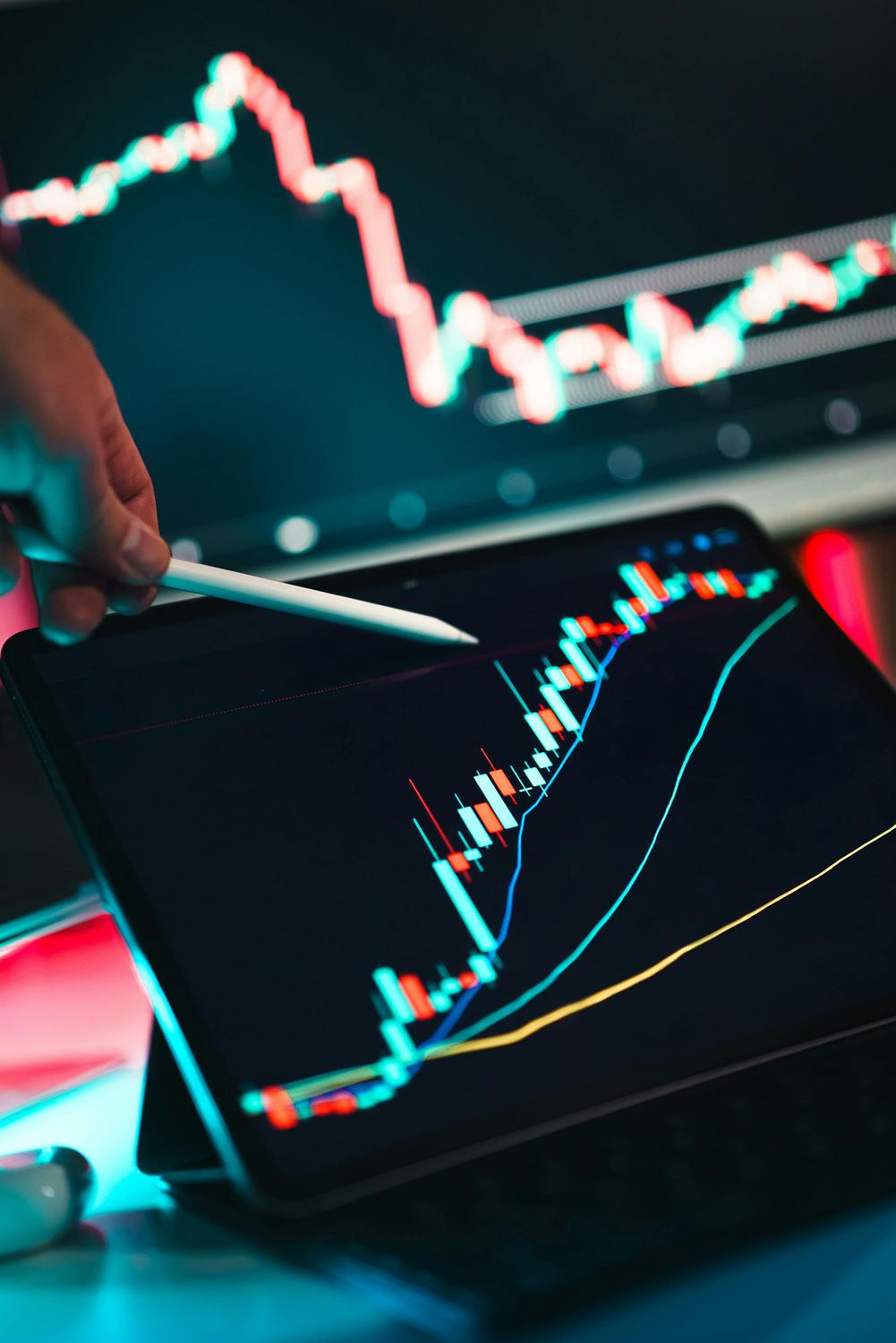 A person points at a digital tablet displaying a colorful candlestick stock chart, with another similar chart visible on a monitor in the background.