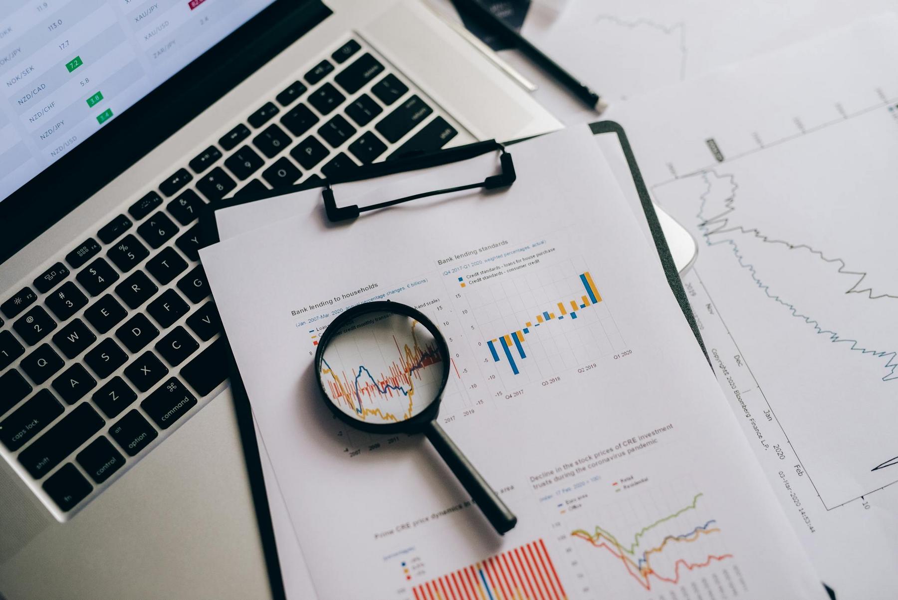 A magnifying glass rests on financial charts and graphs on paper, placed on a laptop keyboard, with data and line graphs visible.
