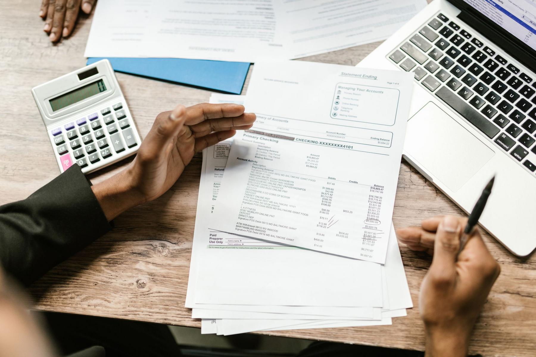 A person holds financial documents at a desk with a calculator, pen, and laptop, appearing to review statements or invoices.