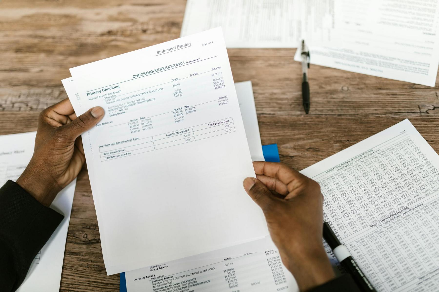 A person holds a checking account bank statement above a wooden table with financial documents, a pen, and a calculator.
