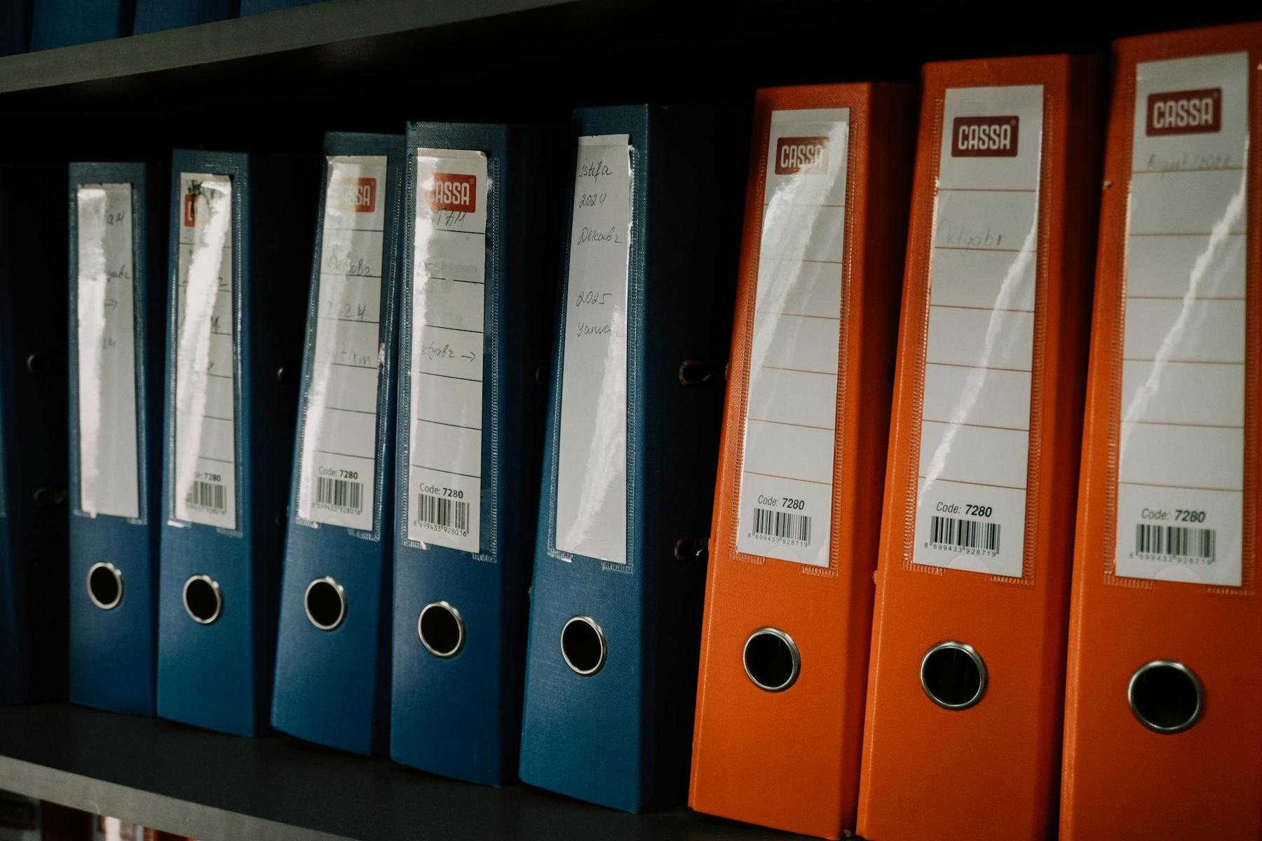 A row of blue and orange ring binders with labels on a shelf, organized vertically and closely packed together.