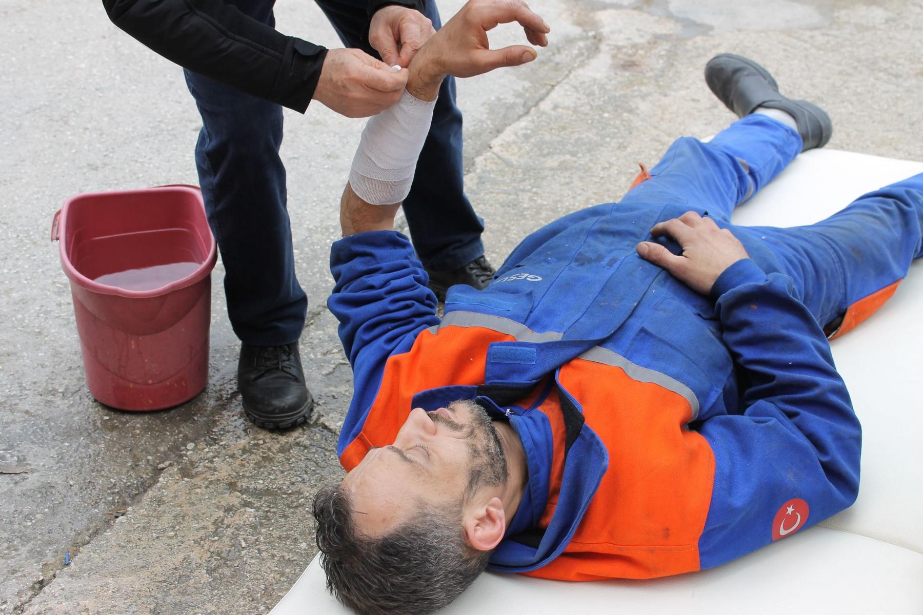 A person in a blue and orange uniform lies on the ground while another person bandages their injured forearm; a red bucket is nearby.