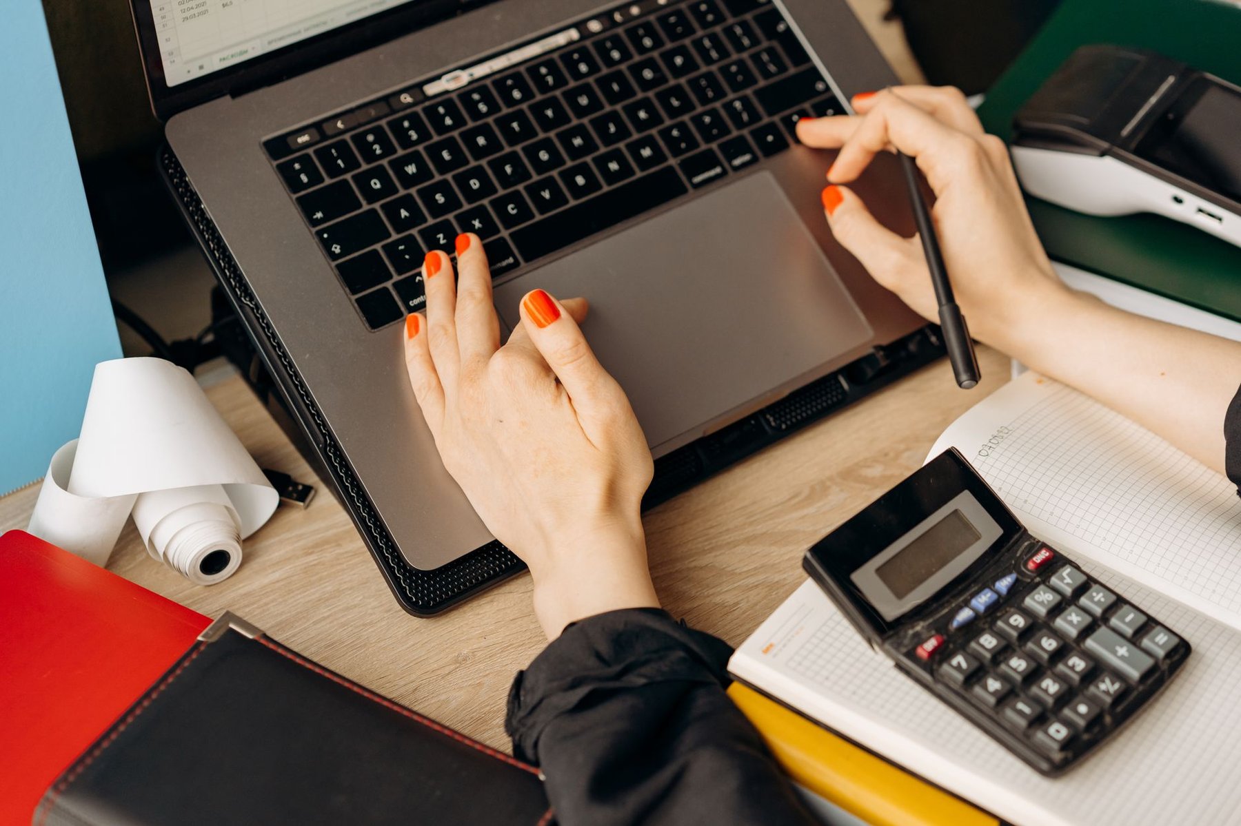 Sole trader doing their bookkeeping on a laptop, with a calculator, notebook, receipt roll and card payment terminal on a wooden desk